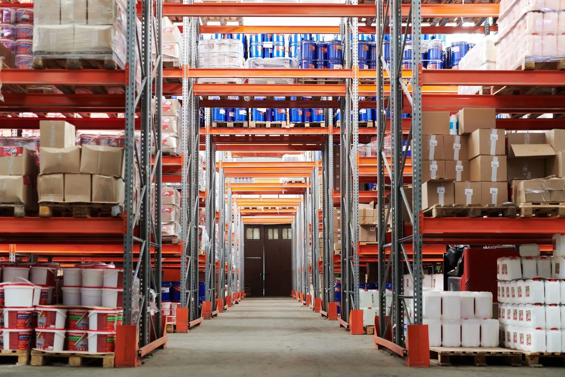 A warehouse aisle between tall orange pallet racking stocked with cartons and drums