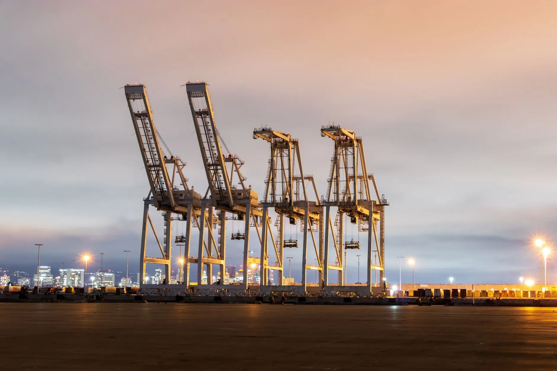Four container cranes lit up at a port under an evening sky