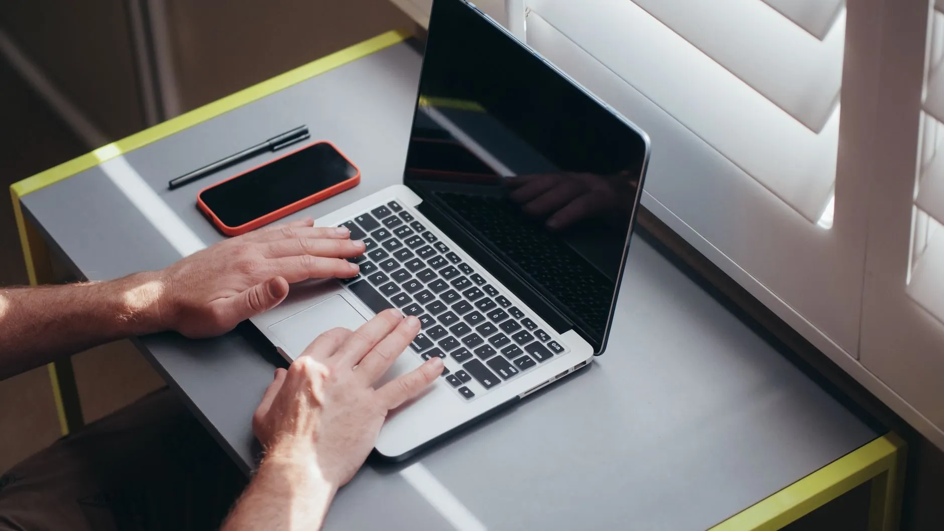 Hands typing on a laptop on a grey desk beside a phone and a pen