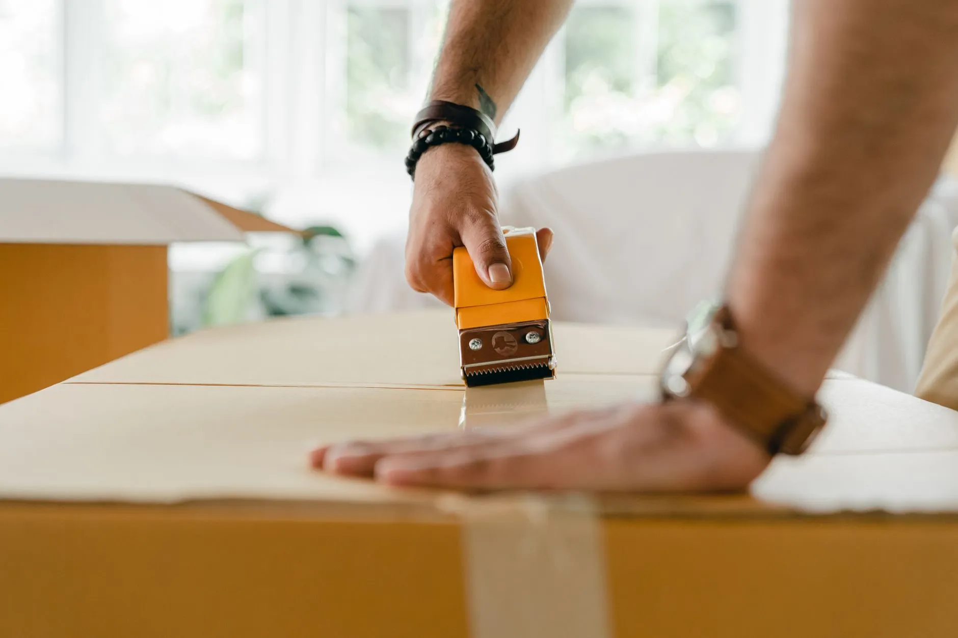 A hand running a tape dispenser along the seam of a large cardboard box